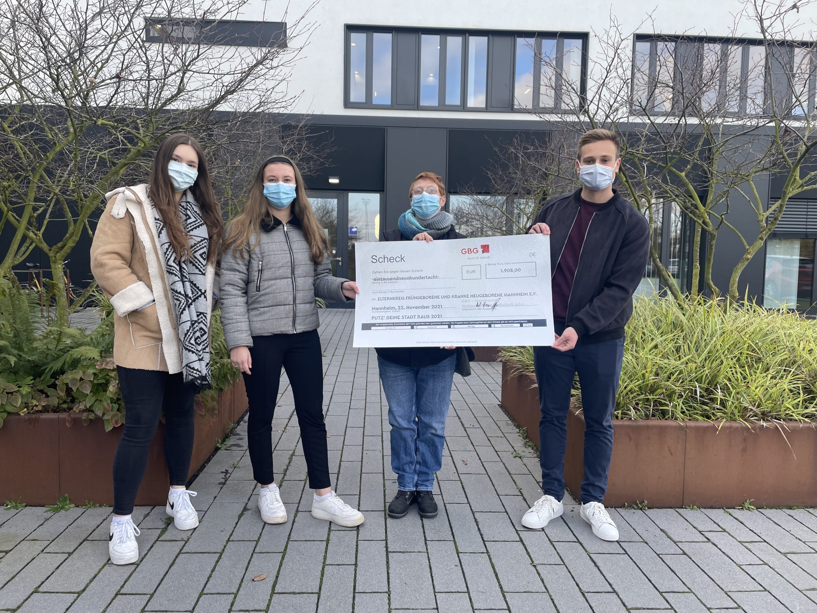 KI-basierte Bildbeschreibung | Vier maskierte Personen stehen draußen auf einem gepflasterten Weg und halten einen großen Scheck vor einem modernen Gebäude zusammen.